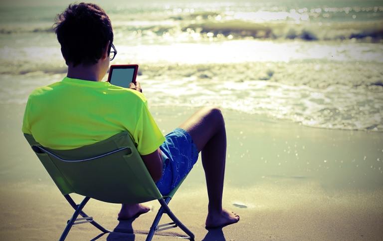Young man with ipad in sun chair, seen from behind on beach by the water  Photo: ChiccoDodiFC