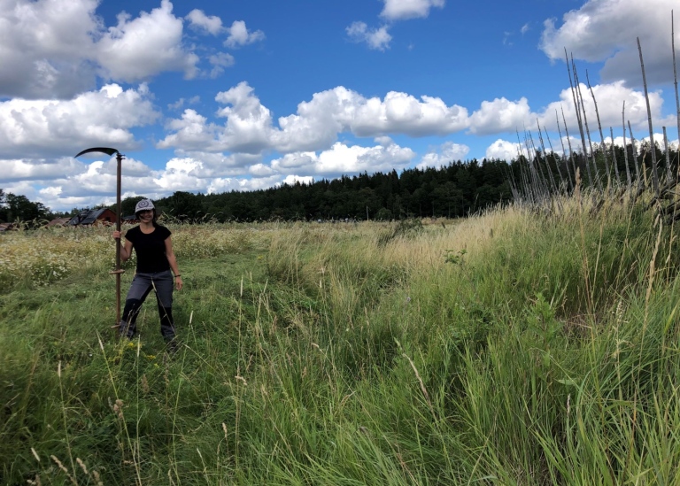 Woman standing in a meadow holding a scythe.