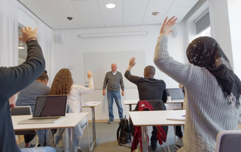 Vuxna elever i ett klassrum räcker upp handen. 