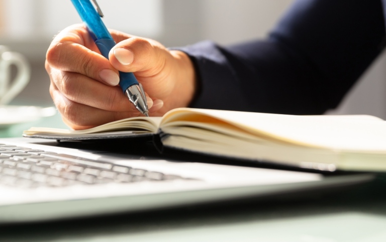 Hand som skriver med penna i block och en laptop. Foto: Andrey Popov, MostPhotos