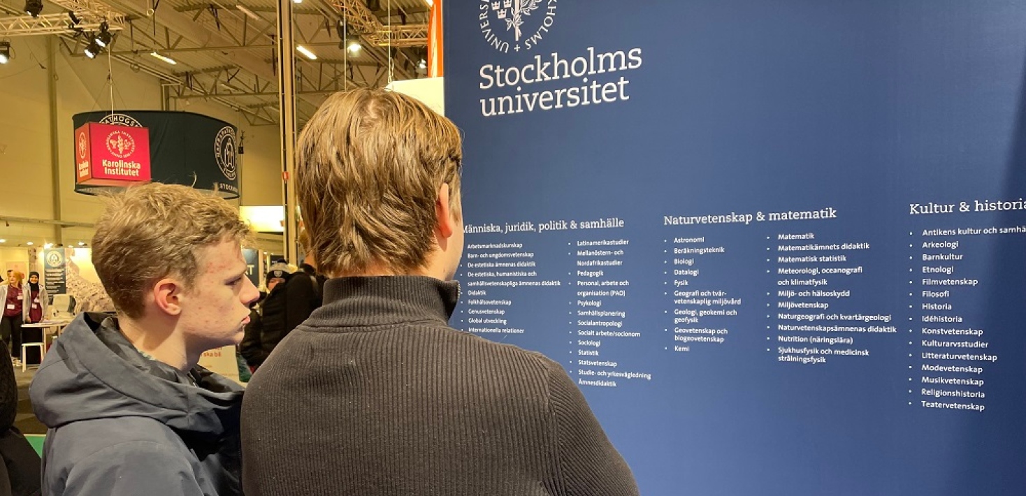 Två killar står och tittar på listan med ämnen på Stockholms universitets montervägg.