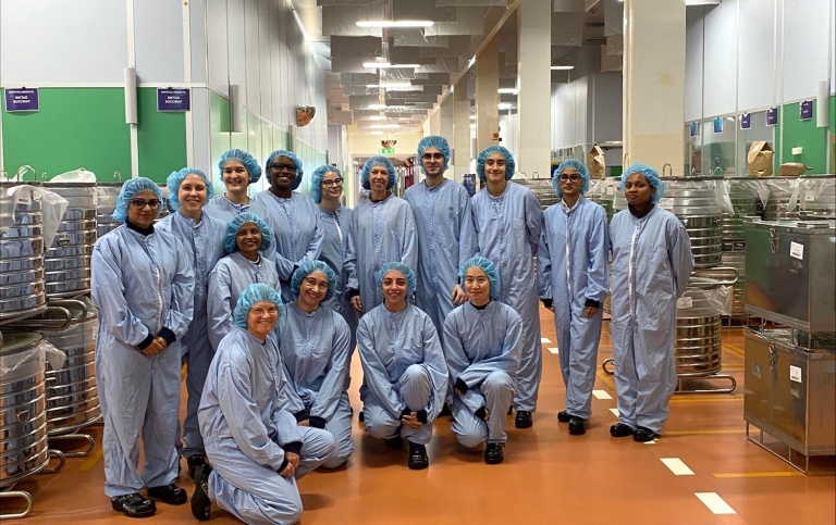 Group photo of 15 people in blue overalls and hairprotection in a large industrial space.