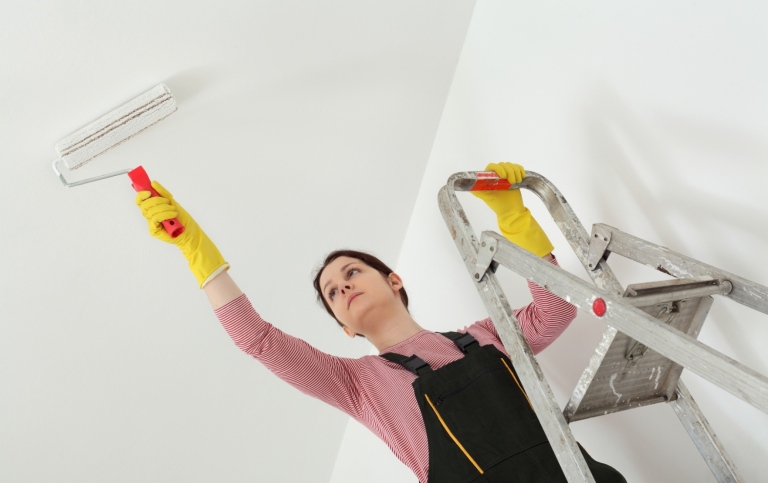 Woman on ladder painting the ceiling with a roller.
