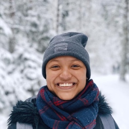 A biracial woman dressed in layers and a beanie, standing outside with a snowy forest background