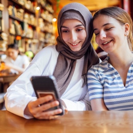 Two happy-looking teenage girls are sitting at a table, looking at the same mobile phone.