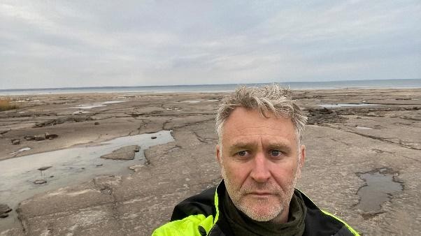 Image of a man, visible from the shoulders up, with short gray hair and facial stubble wearing cold-weather clothes consisting of a bright yellow jacket with a high-collared black shirt. The man is standing in front of a rocky, gray landscape with a body of water in the distance and cloudy skies.