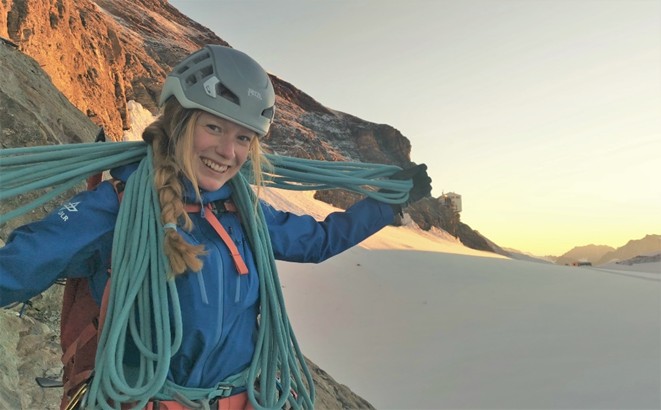Image of a smiling woman, visible from the waist up, with blonde, braided hair wearing a helmet and cold-weather clothes consisting of a blue coat with a high collar. The woman is wearing a backpack and is holding several meters of rope. The woman is standing in front of rock mountains and a snow-covered valley with clear skies.