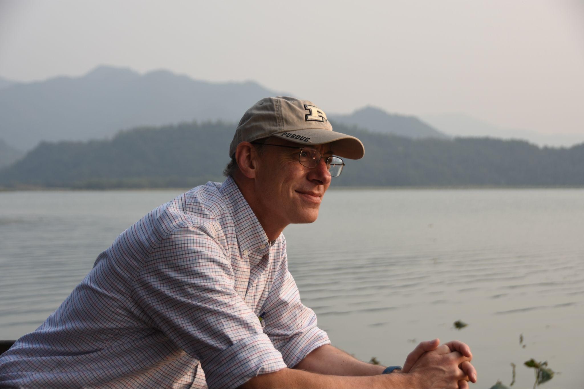 Profile image of a smiling man, visible from the waist up, wearing glasses, a baseball cap and a light gray shirt with the sleeves rolled up to the elbow. The man is leaning on an object, not visible in the image, looking to the right of the image. The man is standing in front of a body of water with mountains and the sky in the background.
