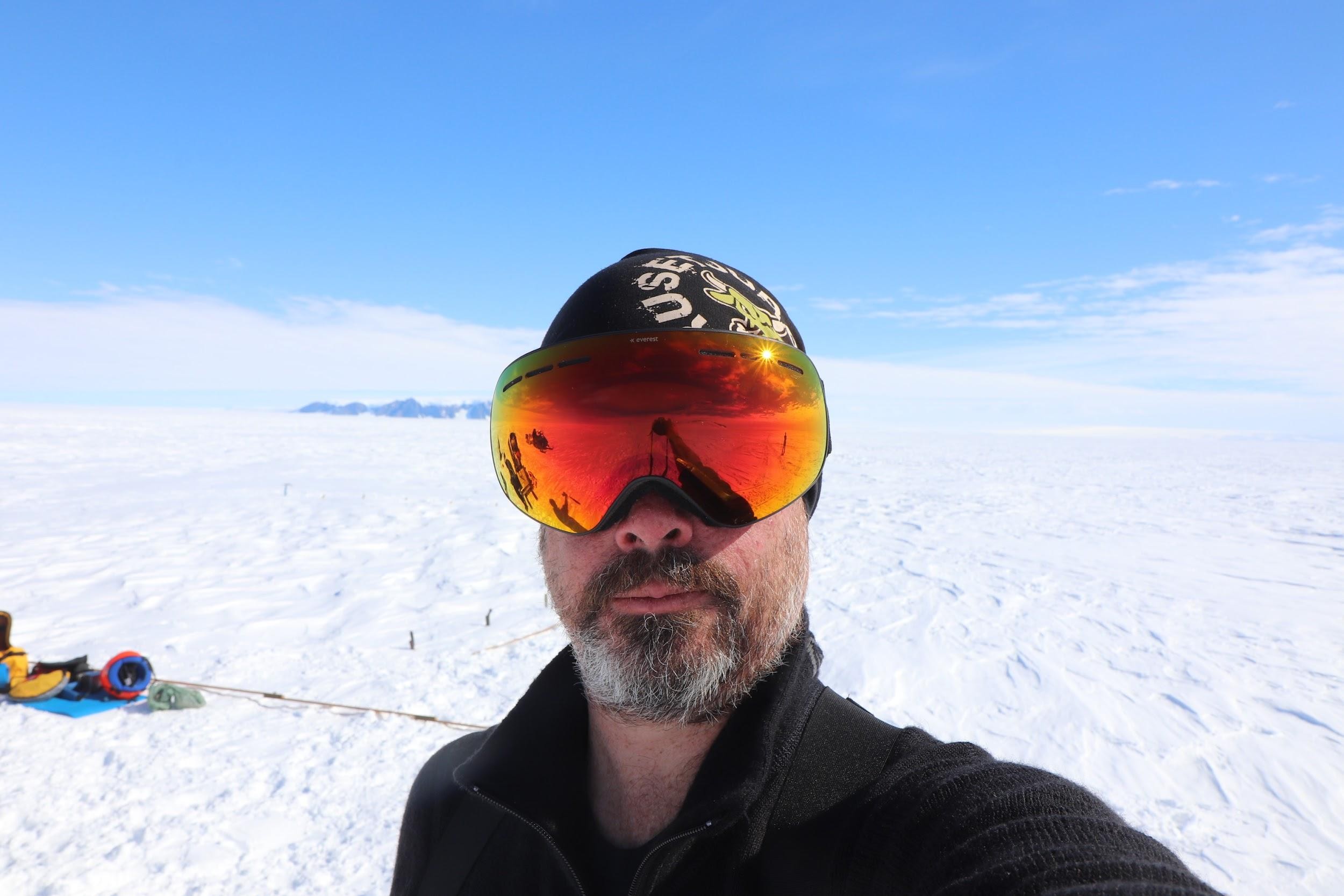 Image of a smirking man, visible from the chest up, with a bead and mustache, wearing a tight-fitting cap on his head along with reflective snow glasses and a black sweatshirt without a hood. The man is standing in front of a snowy landscape with some small rocks in the background and blue skies with some clouds on either side. Behind the man gear is pictured on the ground.