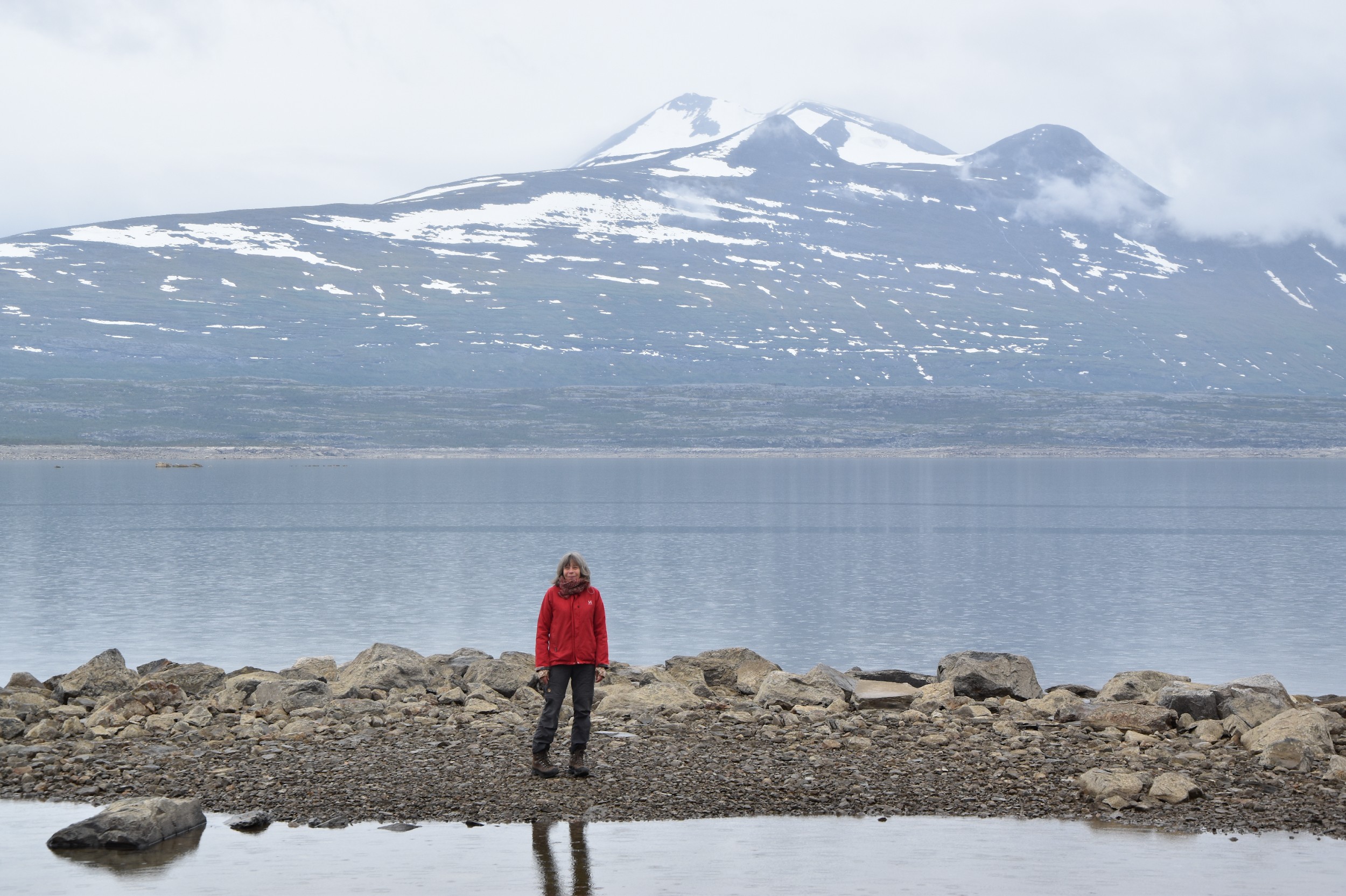 Image of a person, entire body pictured, seen from a distance and wearing cold-weather clothing including a red coat with a hood, gray pants, and boots. The person is seen standing on a rocky area with water in front of and behind the standing location. In the background a larger body of water is visible along with a large, snow-capped mountain and clouds around the peak.
