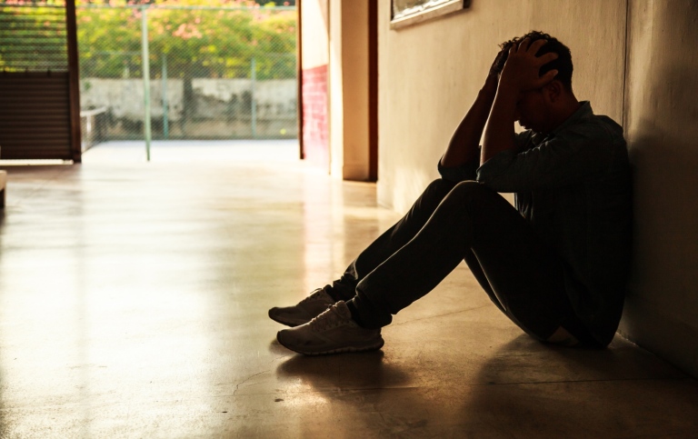 A man sitting on the floor and leaning against the wall. He has his hands in front of his face.