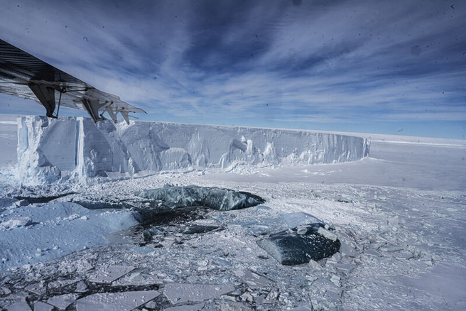 Image of Antarctic ice sheet with dark blue ice, ice shelfs, and ice cliffs visible with an airplane wing.