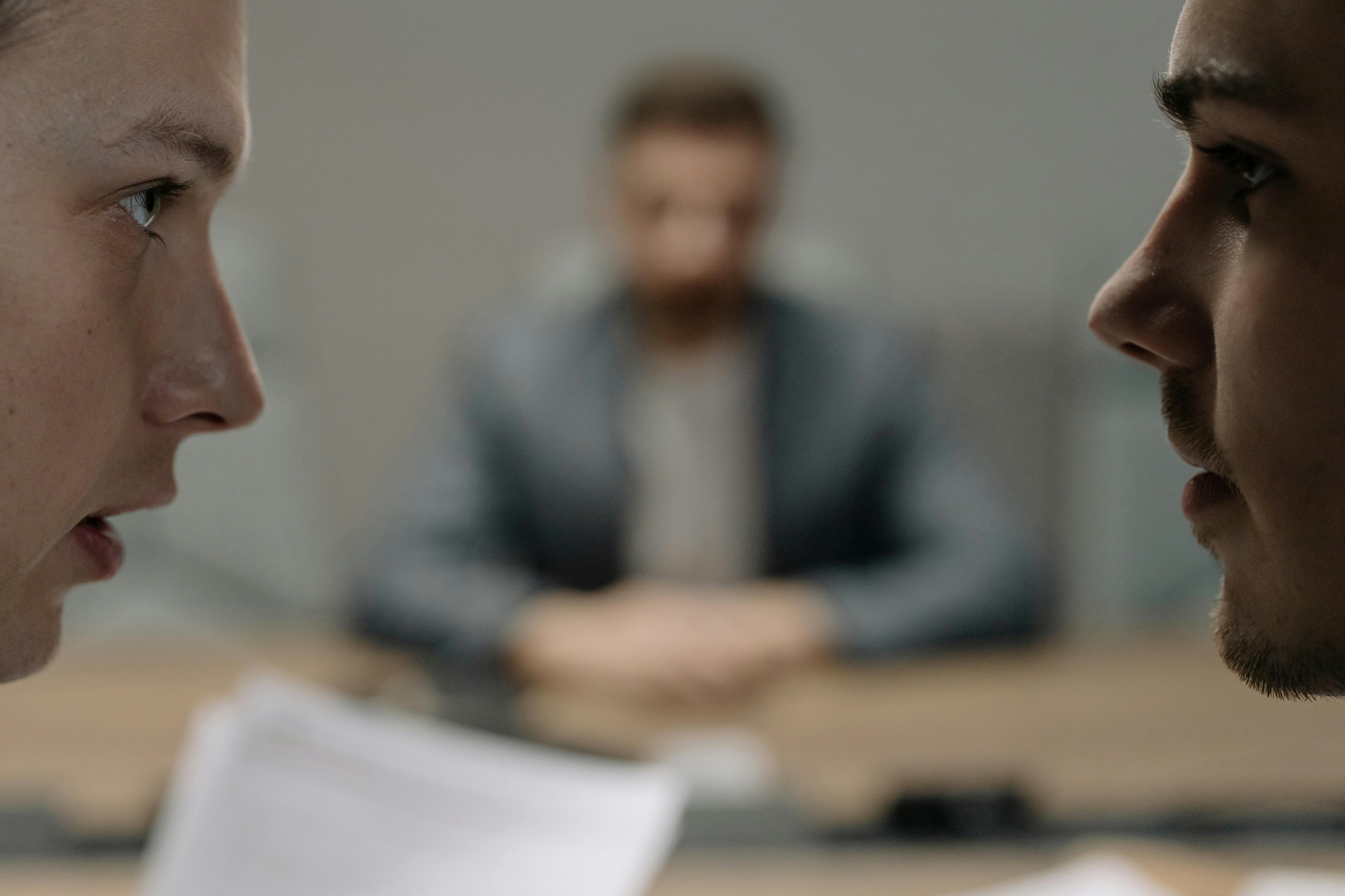 Two people facing each other in the foreground, with a third blurred person in the background.