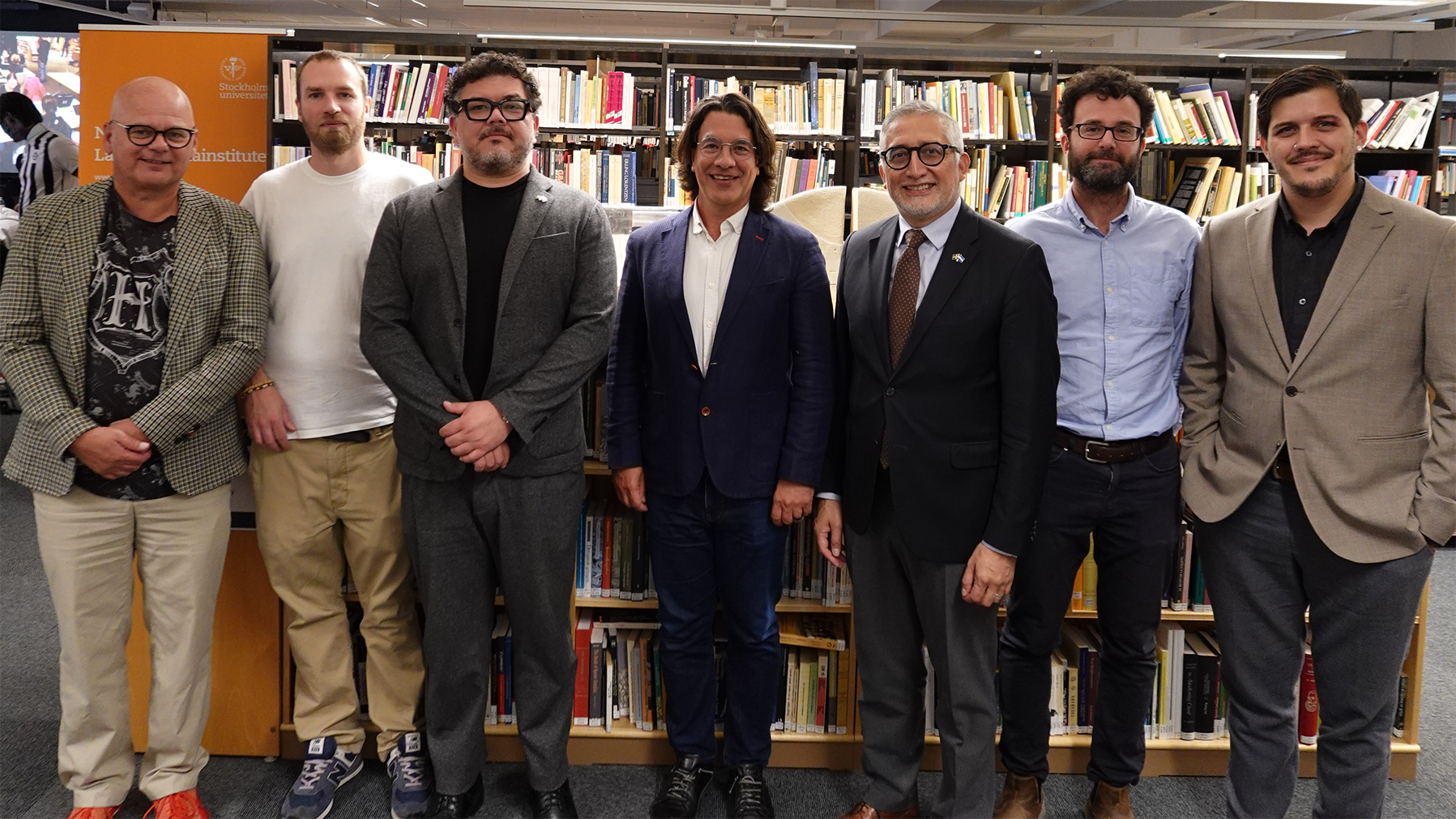 Sju personer står uppställda framför bokhyllor i ett bibliotek eller läsrum. I bakgrunden syns en orange skylt med texten ”Stockholm University” och ”Nordic Latin American Institute”.