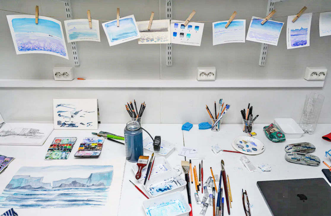 View of an artist’s worktable with watercolor paintings, brushes, paints, and creative materials. Several postcard-sized watercolor artworks hang from a line with clothespins, drying above the workspace.