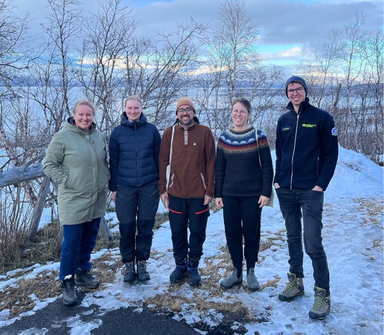 five people standing on some snow, looking at camera, some trees on the background and a lake.