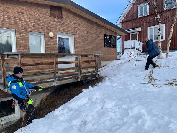 one man pulling another with a rope, during a safety training. they are standing on a slope beside a house, there is some snow on the ground.