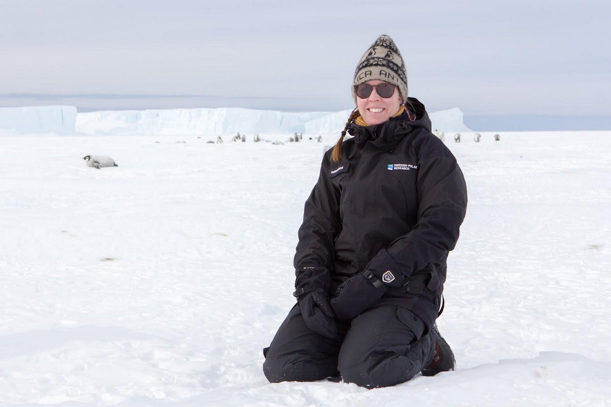 Researcher kneeling on the ice in Antarctica with penguins nearby,.She has sun glasses on and is smilling at the camera.