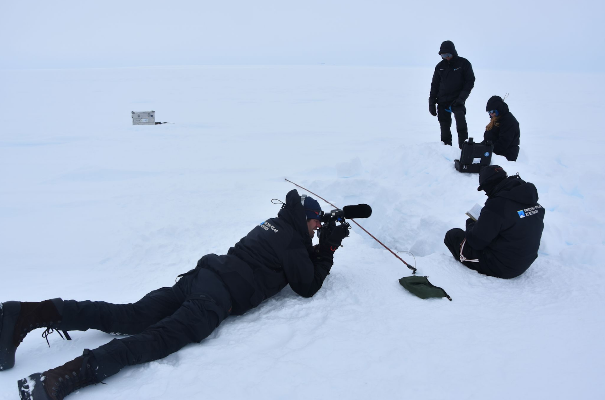 Image of Bram laying on the icy, snow-covered ground while photographing iQ2300 researchers