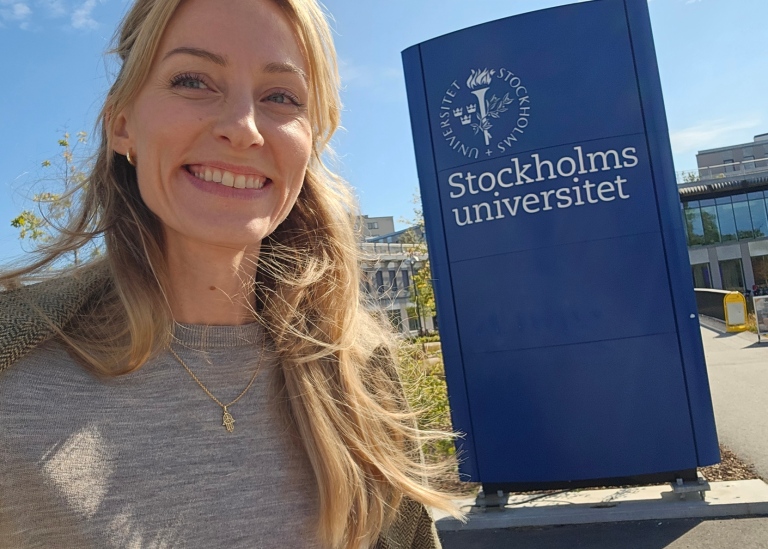 Sofia Lindblom vid blå skylt med Stockholms universitets logotyp framför Campus Albano.