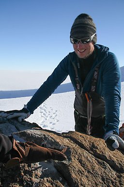 Image of a smiling man, seen from the thighs up, wearing sunglasses and a tight-fitting hat. The man is wearing cold-weather gear including a coat and gloves that are blue and gray in color, along with dark-colored pants. The man is seen leaning on a rock in from of him. Behind the man is snow-covered terrain with some mountain visible and blue skies with some clouds near the mountain.