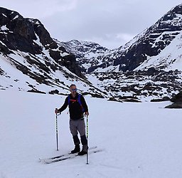 Image of a smiling man on cross-county skis with a ski pole in each hand making contact with the earth. The man has closely-cropped dark hair and dark, short facial hair. He is wearing blue-toned reflective sunglasses, a long-sleeve black shirt, light gray pants, ski boots, and a backpack with blue straps. The man is seen standing in his skis on snow with lightly snow-covered, rocky mountains behind him and a light gray, cloudy sky in the background.