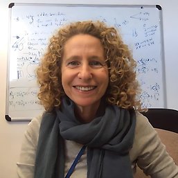 Image of a smiling woman, seen from the chest up. The woman has curly, golden-blond, shoulder length hair. She is wearing a light gray shirt with a dark blue-gray scarf. The woman is seated in front of a whiteboard with calculations appearing behind her in blue pen.