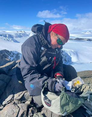 Researcher sitting on some rocks taking notes, with snow in the background, in Antarctica.