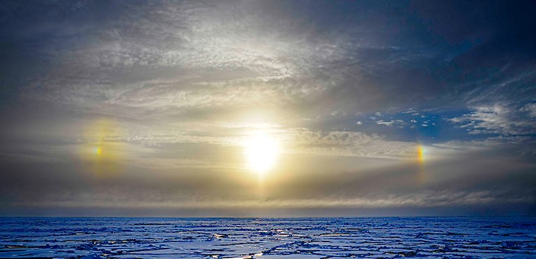 Sol, himmerl med moln, is och hav, två bisolar till höger och vänster.