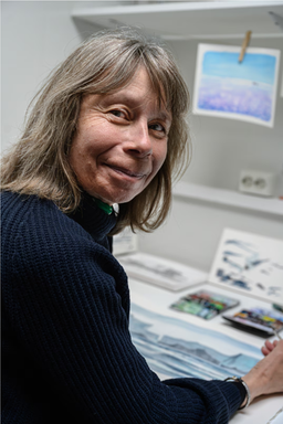 Close-up of an artist smiling at the camera while seated at her worktable. She holds a paintbrush beside a watercolor painting in progress, with art supplies and painting materials visible around her workspace.