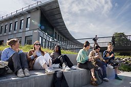 Studenter sitter i solen i Campus Albano, med Hus 2 i bakgrunden.