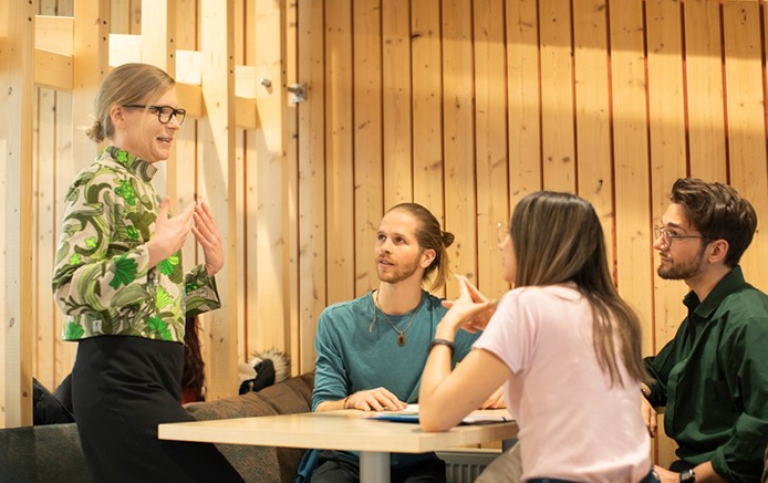 Kvinnlig lärare står vid ett bord och talar med två manliga studenter och en kvinnlig student.