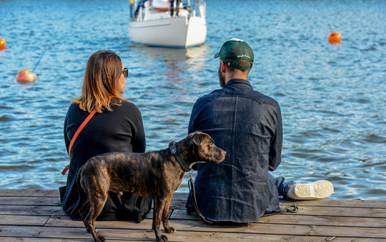  Kvinna och man sitter på bryggkant vid vattnet. En hund står bredvid dem. T@ge Persson / Mostphotos