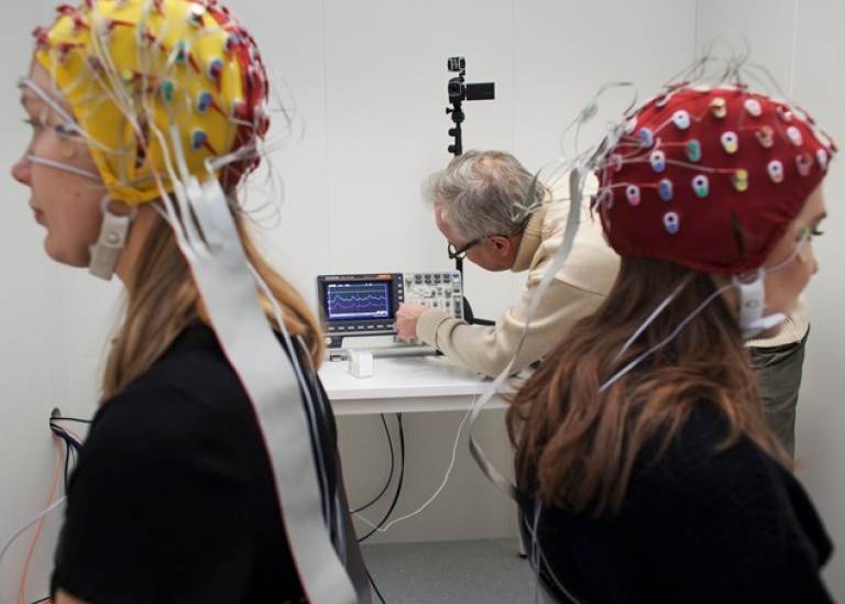 Två studenter med EEG-elektrodmössor på huvudet. I bakgrunden en forskare. Foto: Jens Olof Lasthein