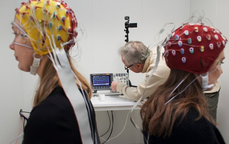 Två studenter med EEG-elektrodmössor på huvudet. I bakgrunden en forskare. Foto: Jens Olof Lasthein