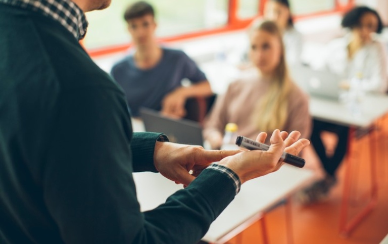 Lärare står inför en klass och håller ett föredrag. Närstudie av händer.