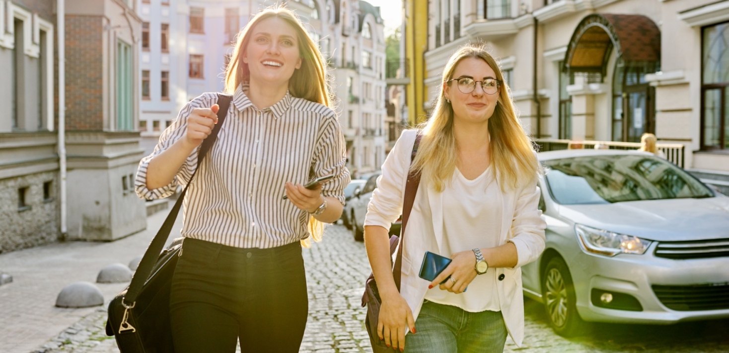 Två kvinnliga glada universitetsstudenter som går längs en statsgata i en europeisk stad.. 