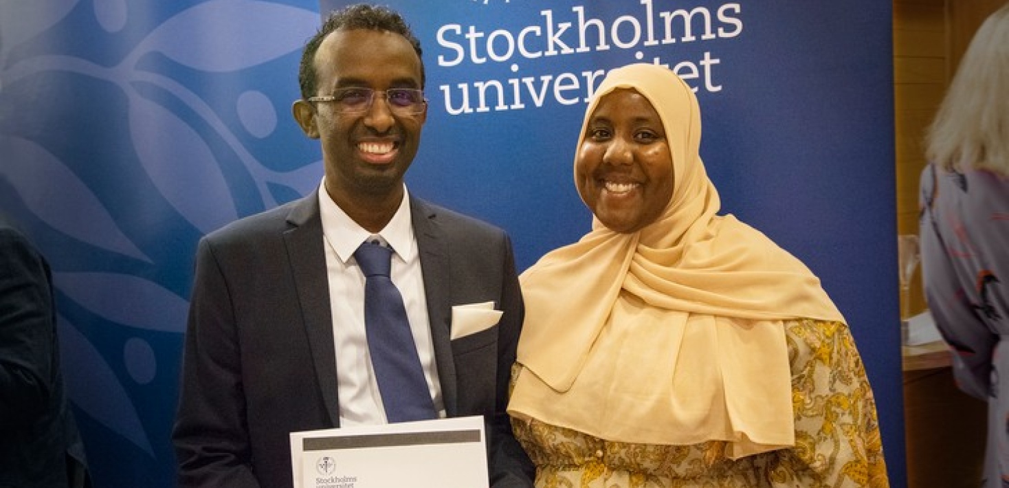 En glad filosofie magister med sällskap, visar diplomet. Kille: kostym, slips. Tjej: klänning, hijab