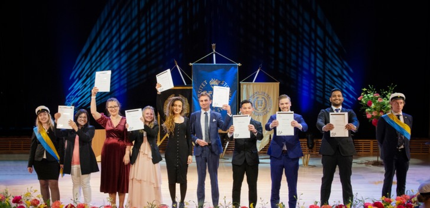 Happy Master of Art students (60 credits) on stage in Aula Magna, waving their diplomas.