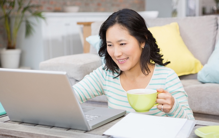 Woman sitting on floor with laptop on table, with mug in her hand. Photo: Wavebreakmedia, MostPhotos