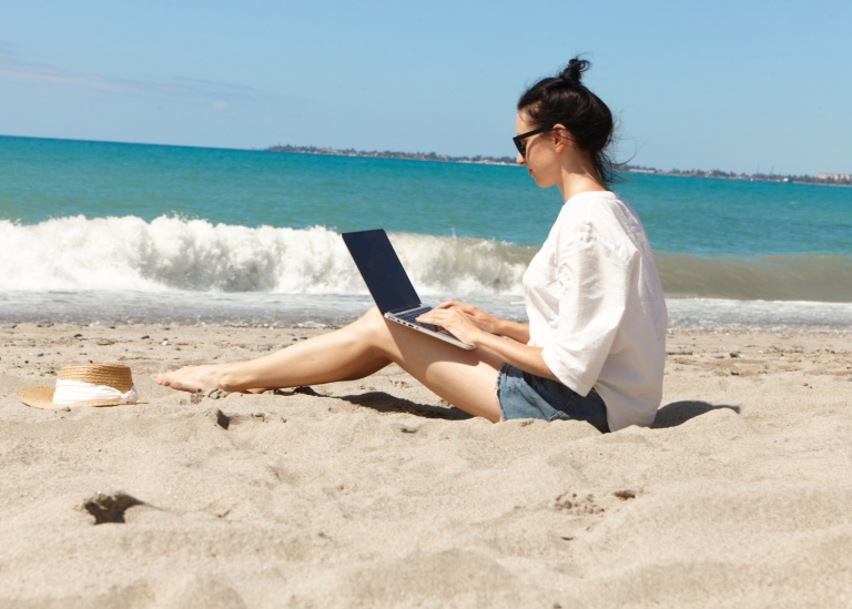 Kvinna sittandes på en strand och skriver på laptop i knät. Foto: Chichina Maria/Mostphotos.