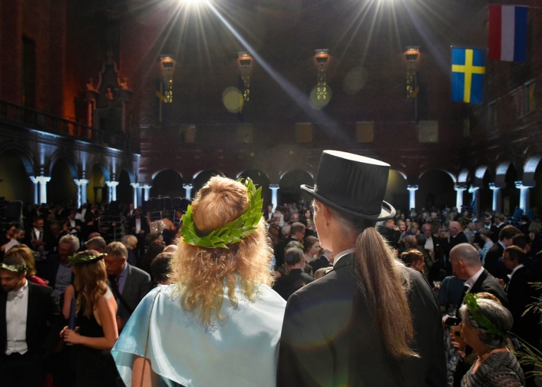 Person with a laurel wreath and  person with a doctoral hat looks out over the Blue Hall in Stockholms City Hall in 2019