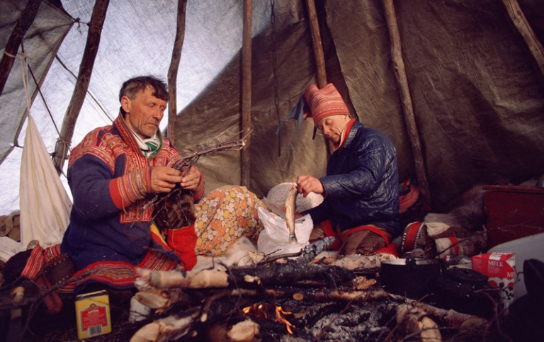 Två samer i en tältkåta som lagar mat över eld.
