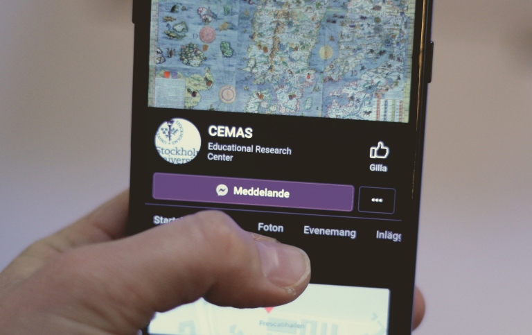 En hand håller i en telefon. På telefonens skärm syns CEMAS facebooksida. 