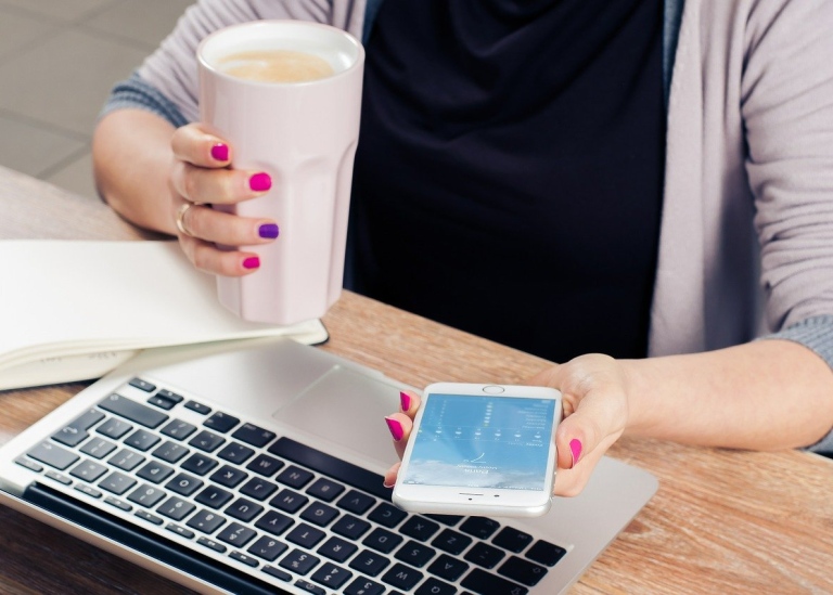 Person at desk with laptop, mobile, notepad and a mug of latte. Photo: Firmbee från Pixabay.