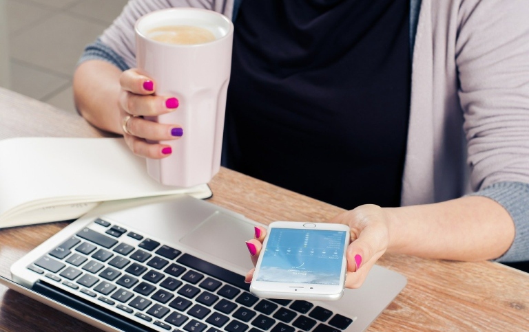 Person at desk with laptop, mobile, notepad and a mug of latte. Photo: Firmbee från Pixabay.