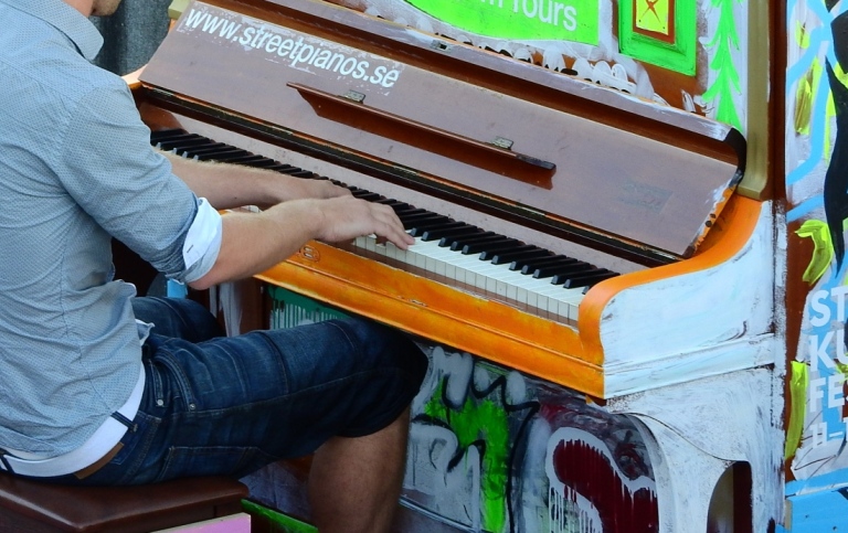En person i grå skjorta spelar på ett piano målat i starka färger. Utomhus.