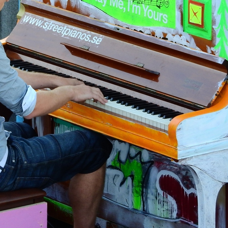 En person i grå skjorta spelar på ett piano målat i starka färger. Utomhus. 