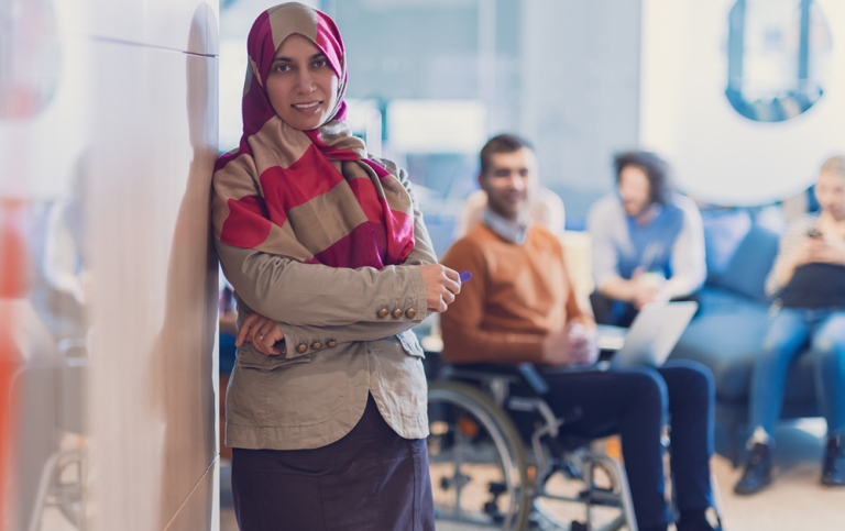 Kvinna med hijab, man i rullstol,personer sitter i en soffa i bakgrunden.