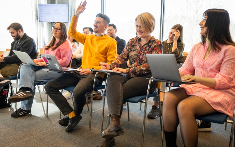 Students sitting on chairs in rows, one holding his hand in the air.